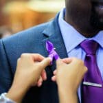 Person pins a purple ribbon on a man's suit jacket.