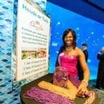 A woman in a pink dress stands next to a colorful craft display and informational board.