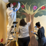 People painting colorful tree mural indoors.